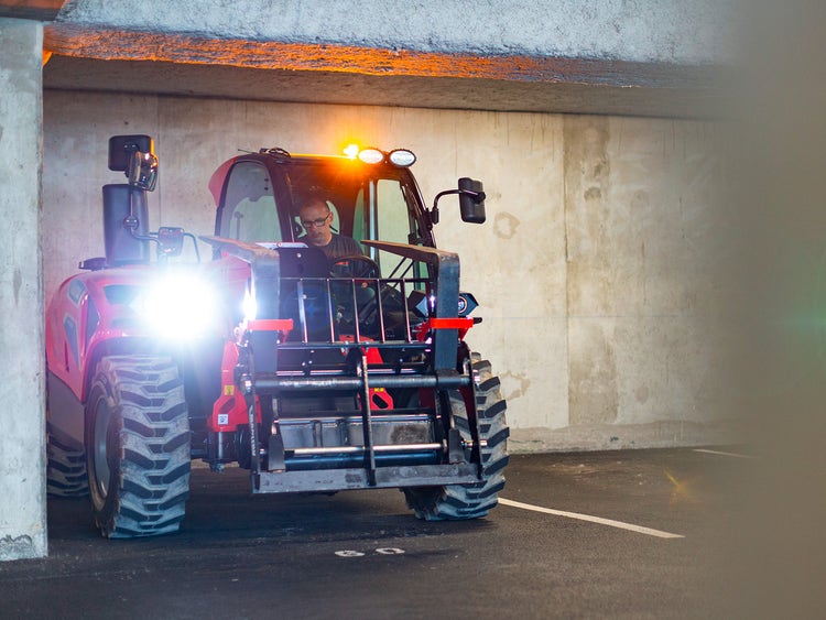 Teleskoplader mit eingeschaltetem Scheinwerfer in einem Parkhaus, ein Mann sitzt in der Fahrerkabine.