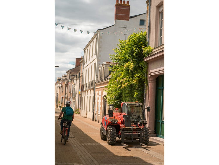 eleskoplader fährt durch eine enge Altstadtgasse, im Vordergrund eine Radfahrerin mit Rucksack.