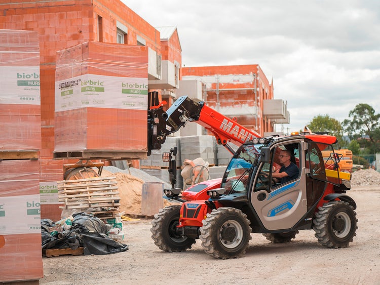 Manitou Teleskoplader hebt auf einer Baustelle eine Palette mit Ziegelsteinen, Fahrer sitzt in der Kabine.
